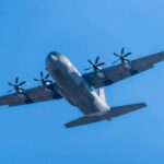 A C-130 Hercules military aircraft soaring in clear blue skies.